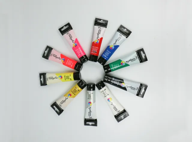 Twelve System 3 acrylic paint tubes arranged in a circular pattern on a white background