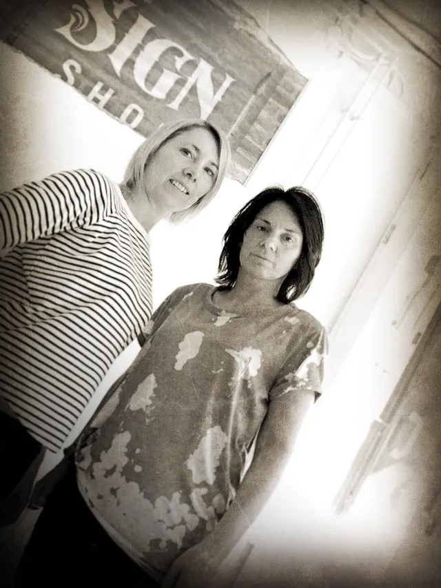 Two women standing in front of rustic wooden signboard reading 'Sign Shop'