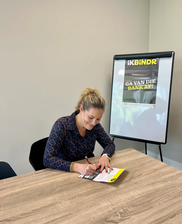 Vrouw ondertekent document aan tafel met IK BINDR-promotieposter op achtergrond