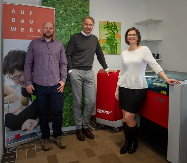 Three people standing next to Trotec Speedy series CO₂ laser cutter in office setting