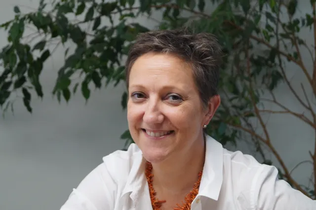Glimlachende vrouw in wit shirt met oranje halsketting, zittend binnen bij groene bladplanten
